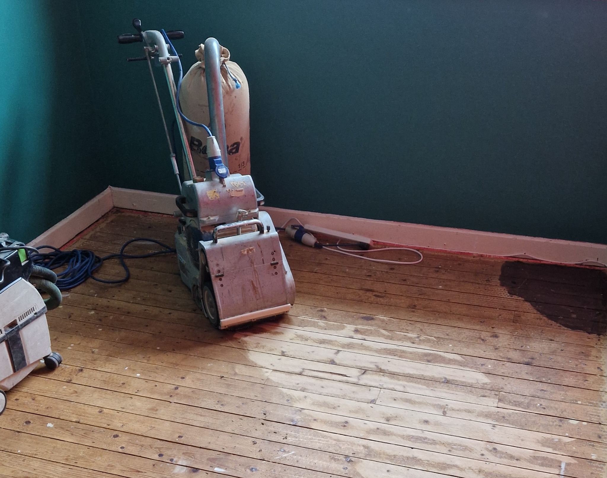 Eco-Friendly Wood Floor Sanding in London – Isleworth Victorian pine floorboards mid-restoration with a low-dust belt sander in a teal-walled bedroom.