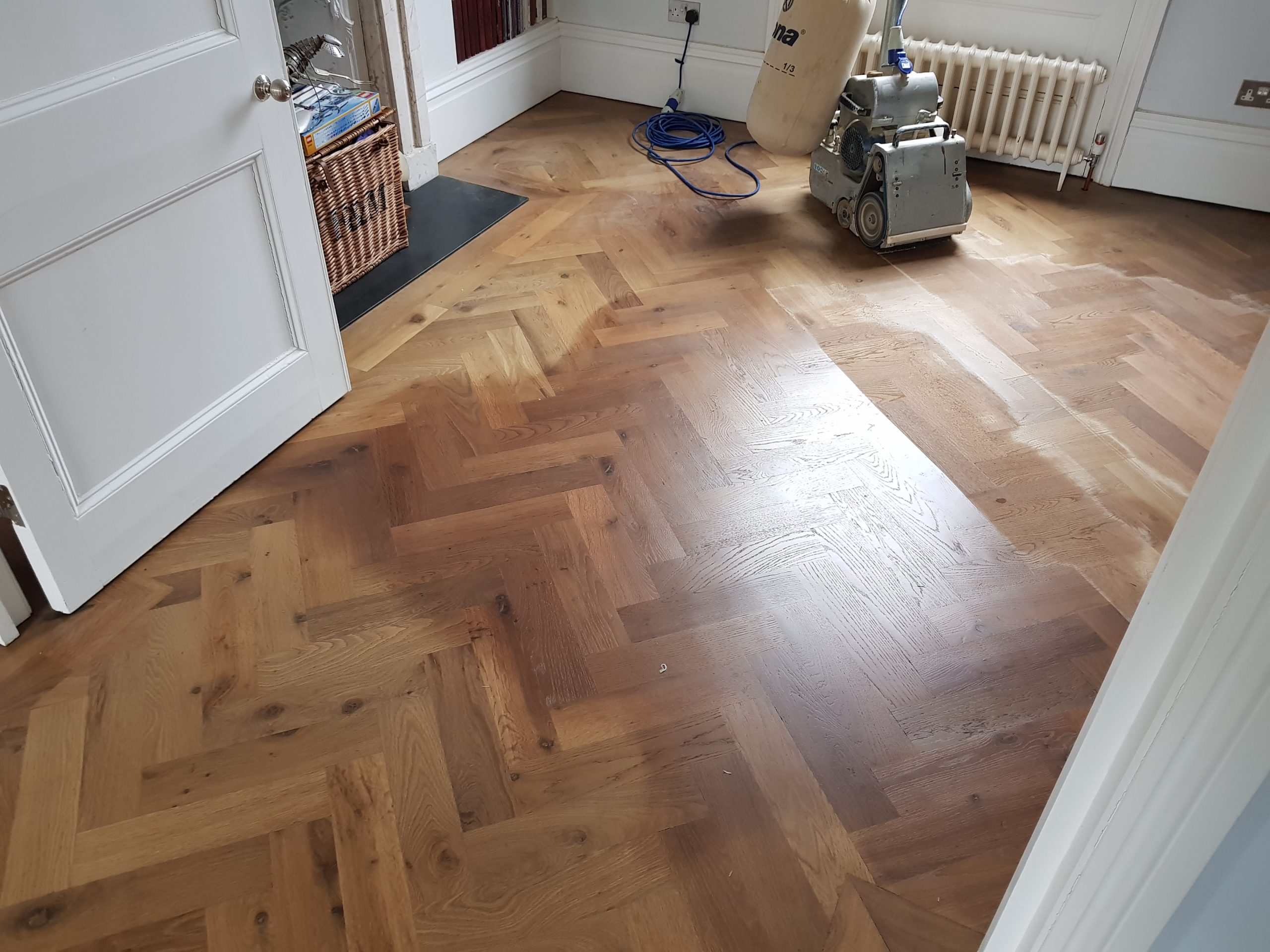 Heritage Parquet sanding in Isleworth — oak herringbone parquet wood floor mid-restoration with a drum sander, showing a half-sanded section and warm grain.