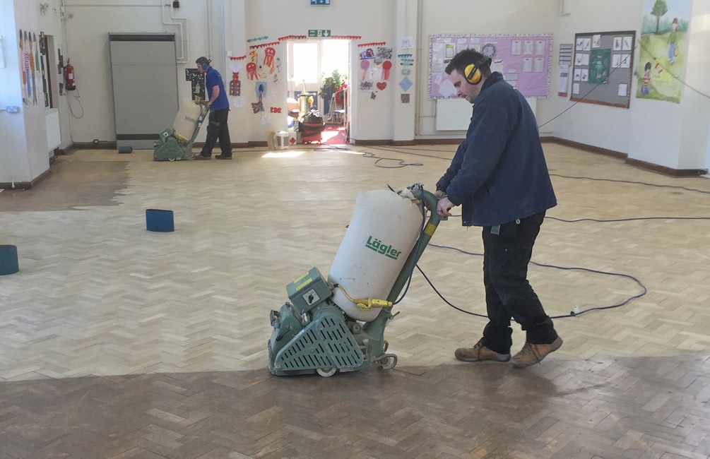 Parquet Floor Sanding in Isleworth school hall, with technicians sanding an oak herringbone parquet floor using professional drum sanders.
