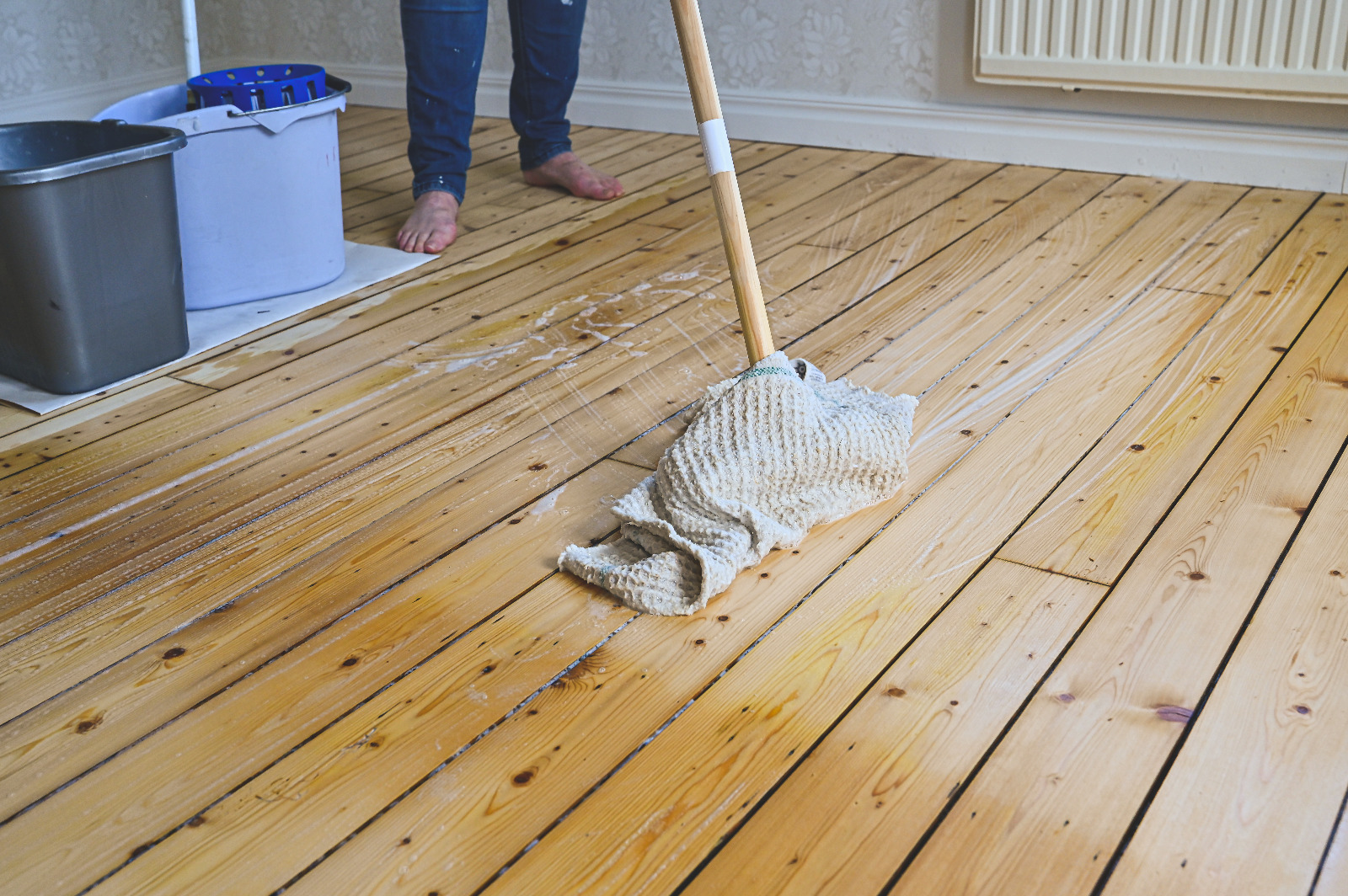 After Wood Floor Sanding in an Isleworth living room, a person is cleaning freshly sanded softwood pine floorboards with a mop and bucket to keep the timber looking its best.
