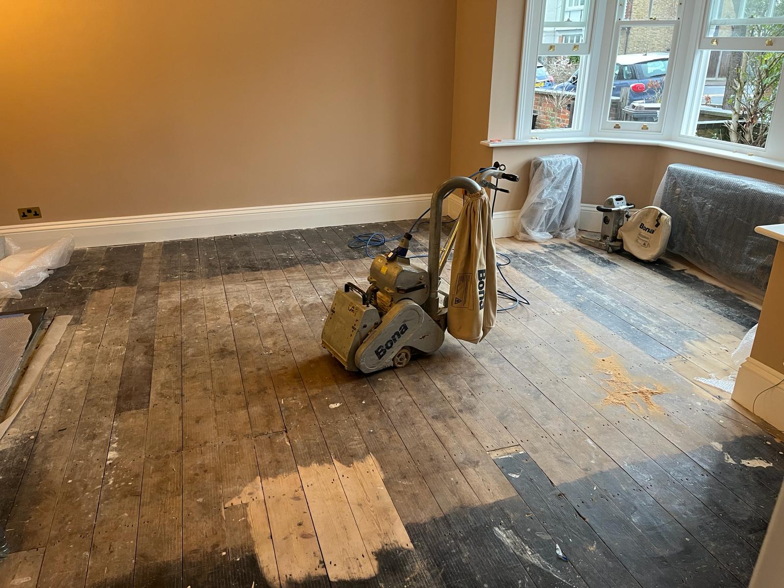 Wood Floor Sanding in Isleworth – Original Pine Floorboards in Progress Wood Floor Sanding in Isleworth living room, showing original pine softwood floorboards mid-sanding with Bona machines removing dark, worn finishes.